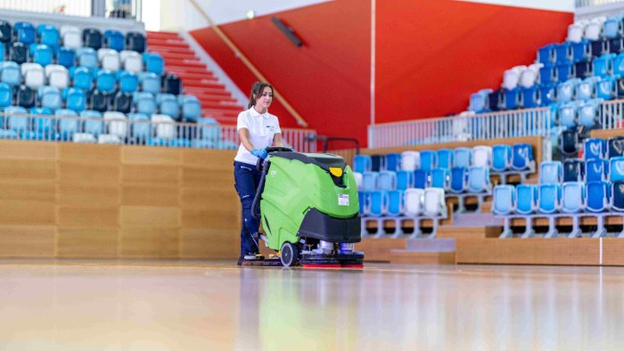 Grüne, große Reinigungsmaschiene für Fußböden, welche von einer Frau durch eine Thurnhalle geschoben wird